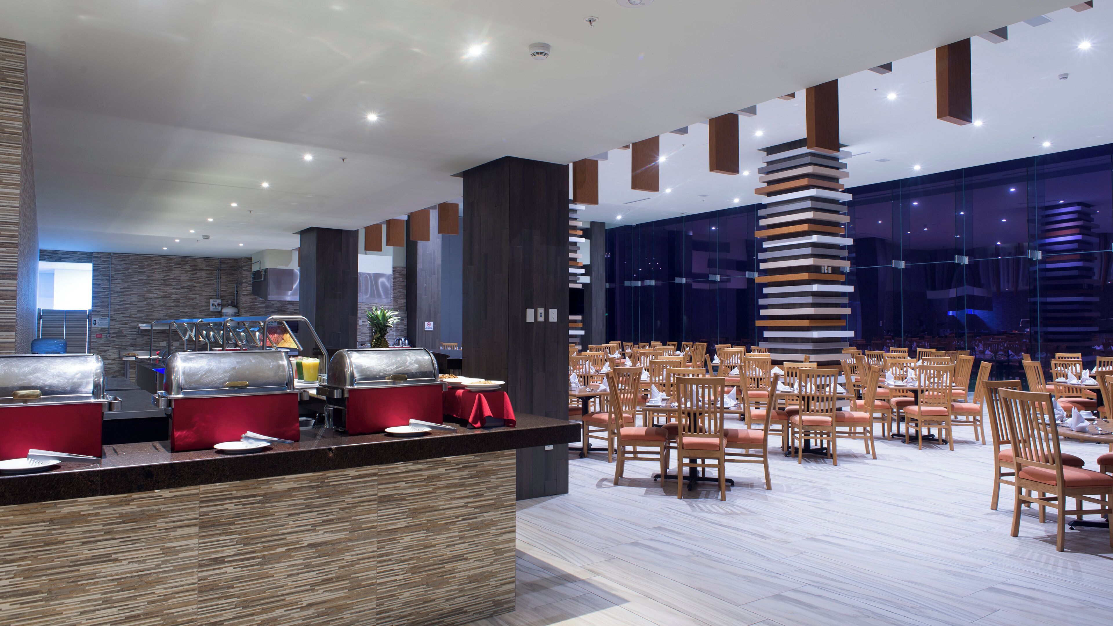 Un espace buffet avec plusieurs présentoirs de nourriture, un coin repas avec des tables et des chaises, et un aménagement intérieur moderne agrémenté d’éléments en bois et d’une grande baie vitrée.