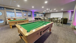 A pool table with a green felt surface, a pool cue, and a billiard ball. There are two pool tables, chairs, and a projector screen in the room. - Yarnfield Park Training Centre (Stone)