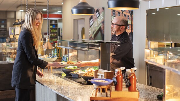 A counter with a variety of food items and condiments, a person serving, and a modern interior design. - Yarnfield Park Training Centre (Stone)