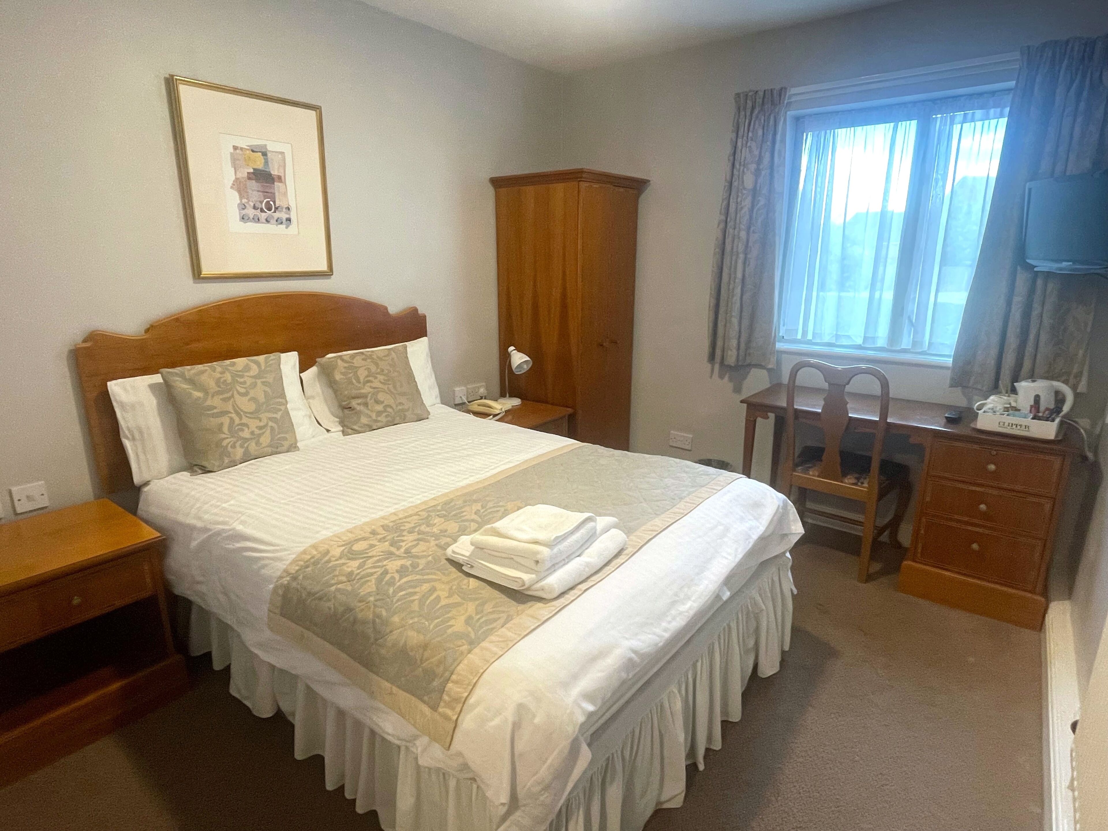 A hotel room with a large bed, a wooden wardrobe, a desk with a chair, and a window with curtains.