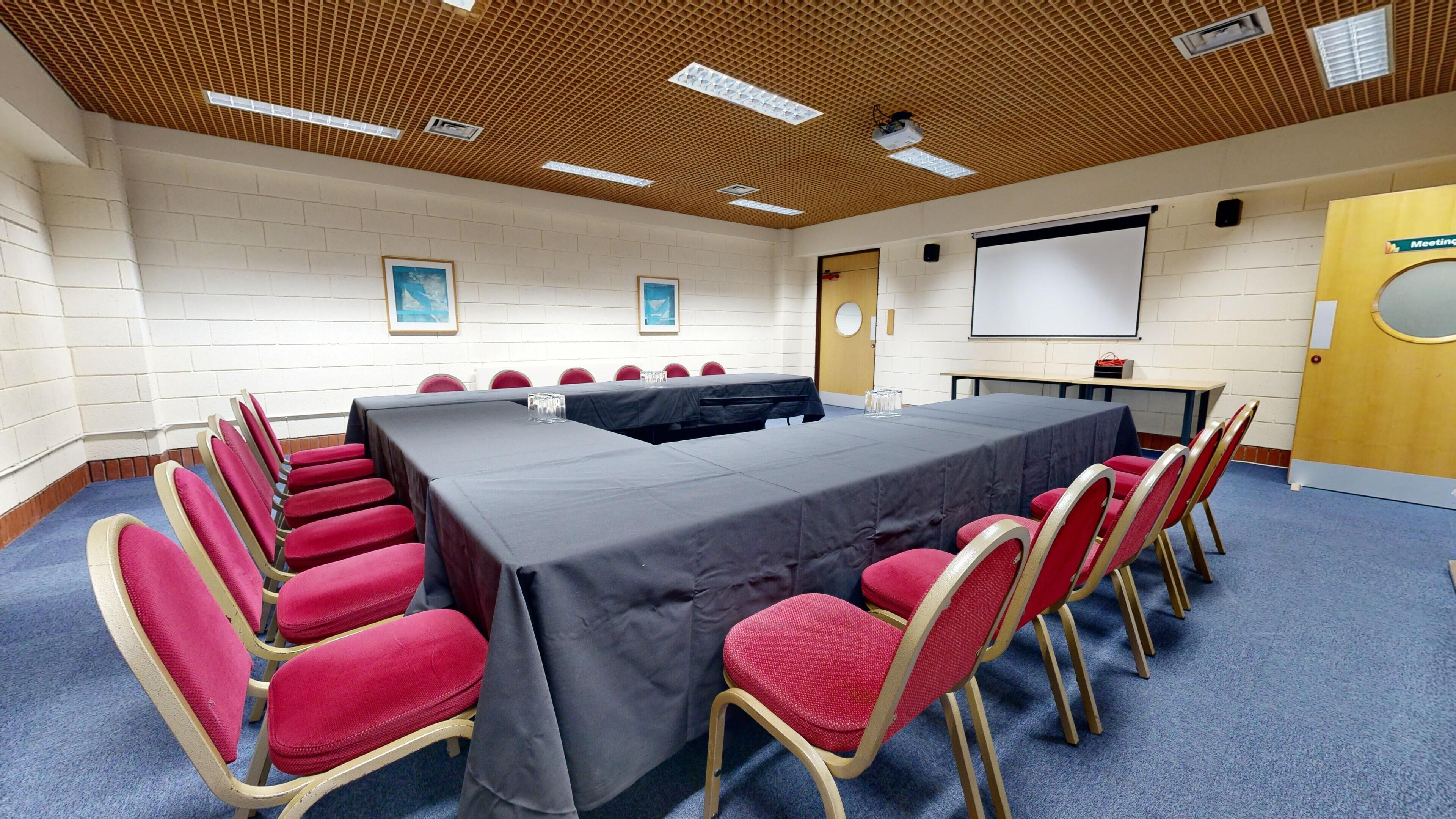A conference room with a long table, red chairs, a projector screen, and framed pictures on the walls.