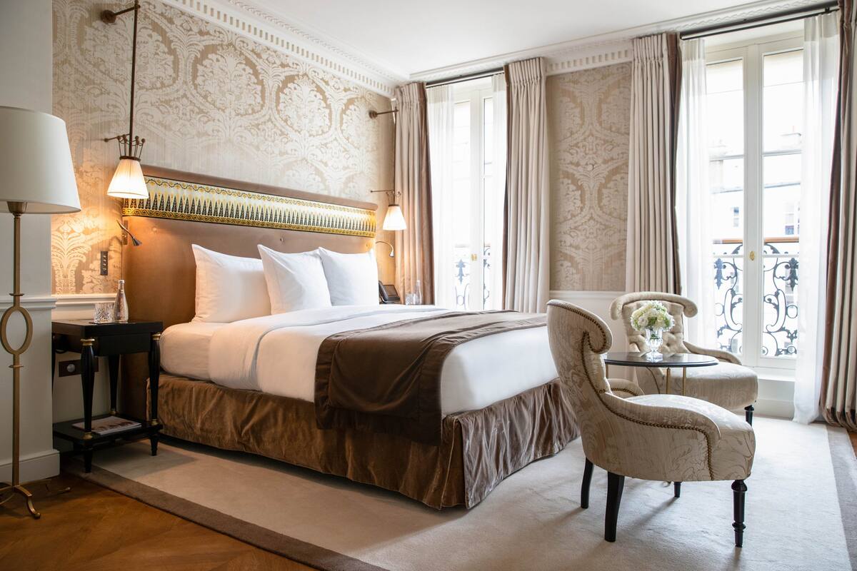 A hotel room with a large bed, two bedside lamps, a small table with a vase, and a chair.