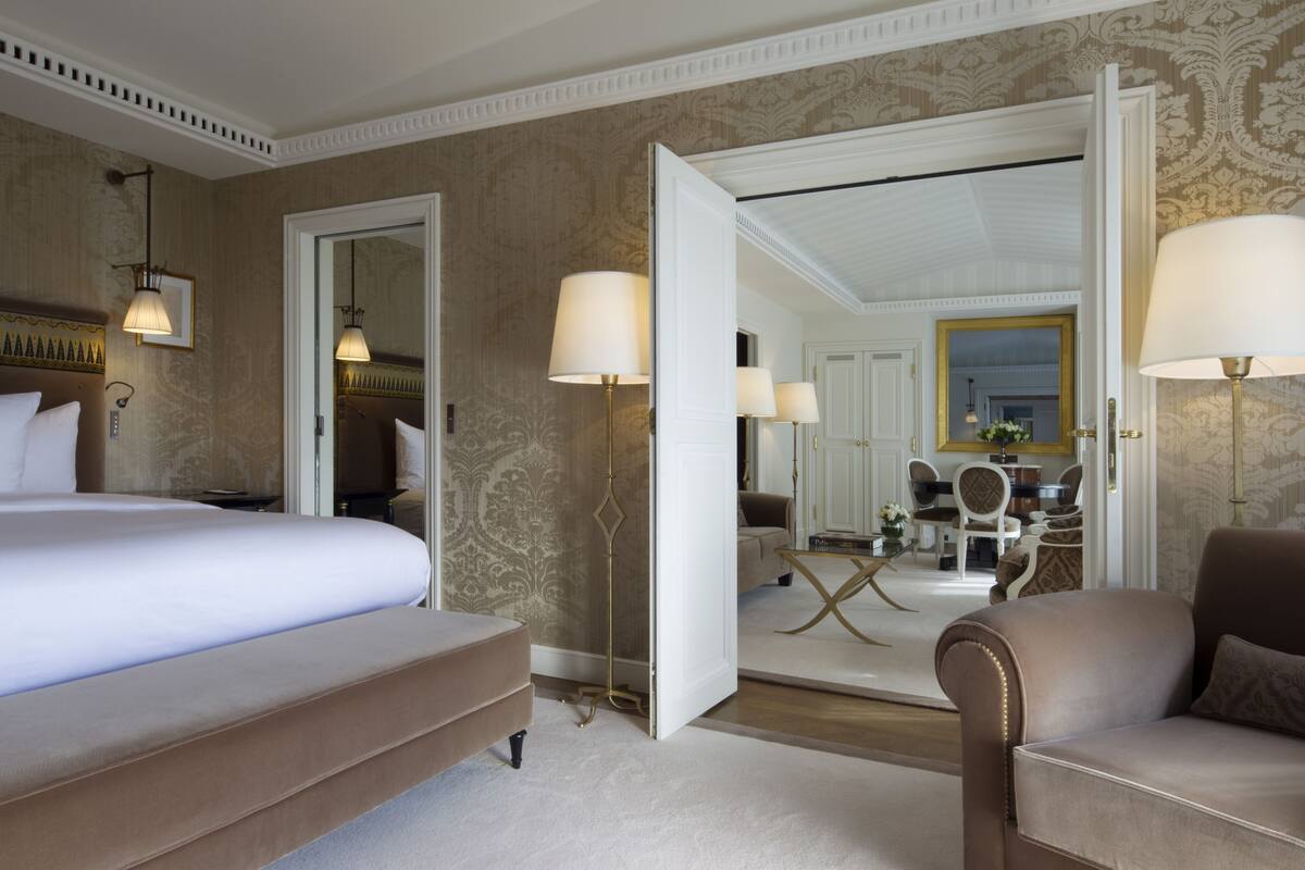 A hotel room with a large bed, a sitting area with a sofa, a dining table, and a mirror reflecting the room.
