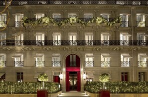 Fachada imponente de un hotel con balcones ornamentales, una entrada central con puertas rojas e iluminación decorativa.
