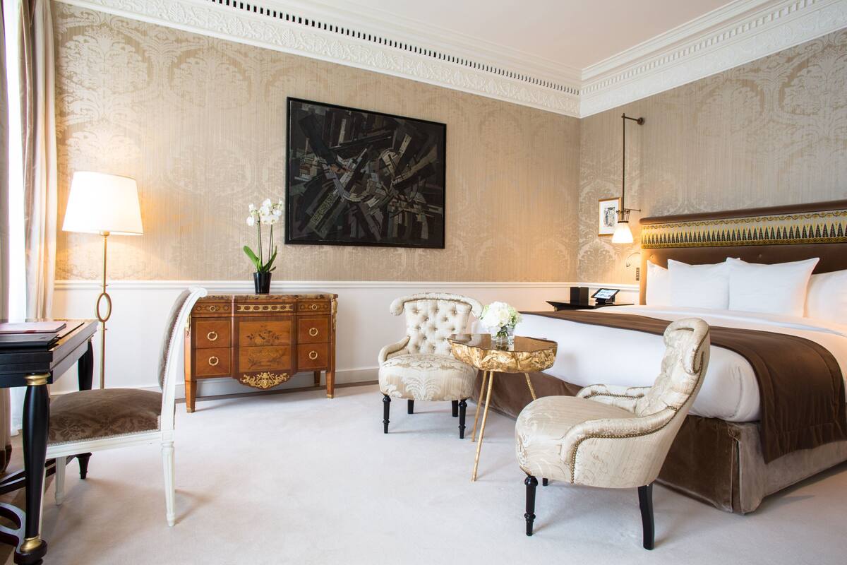 A hotel room with a large bed, two armchairs, a small round table, a dresser, and a painting on the wall.