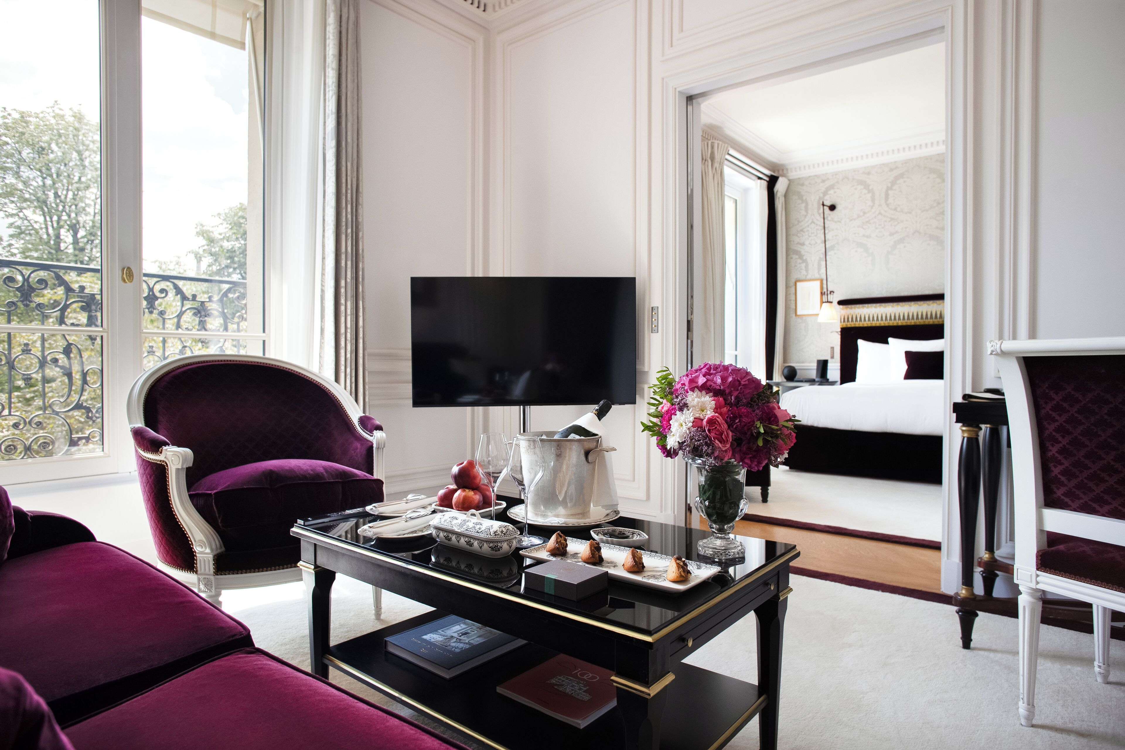 Una lujosa habitación de hotel con una cama grande, televisión, una mesa de comedor con un florero y un balcón con barandilla.
