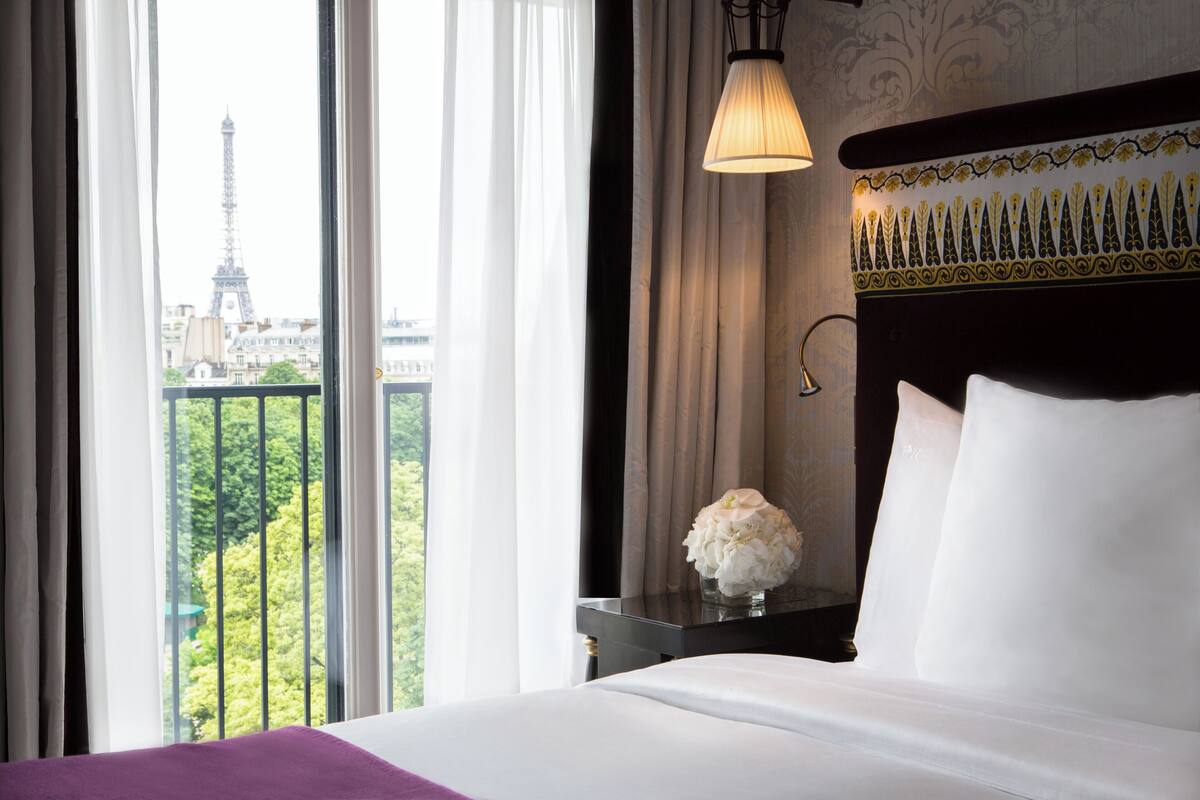 A hotel room with a bed, a nightstand with a vase of flowers, a lamp, and a view of the Eiffel Tower.