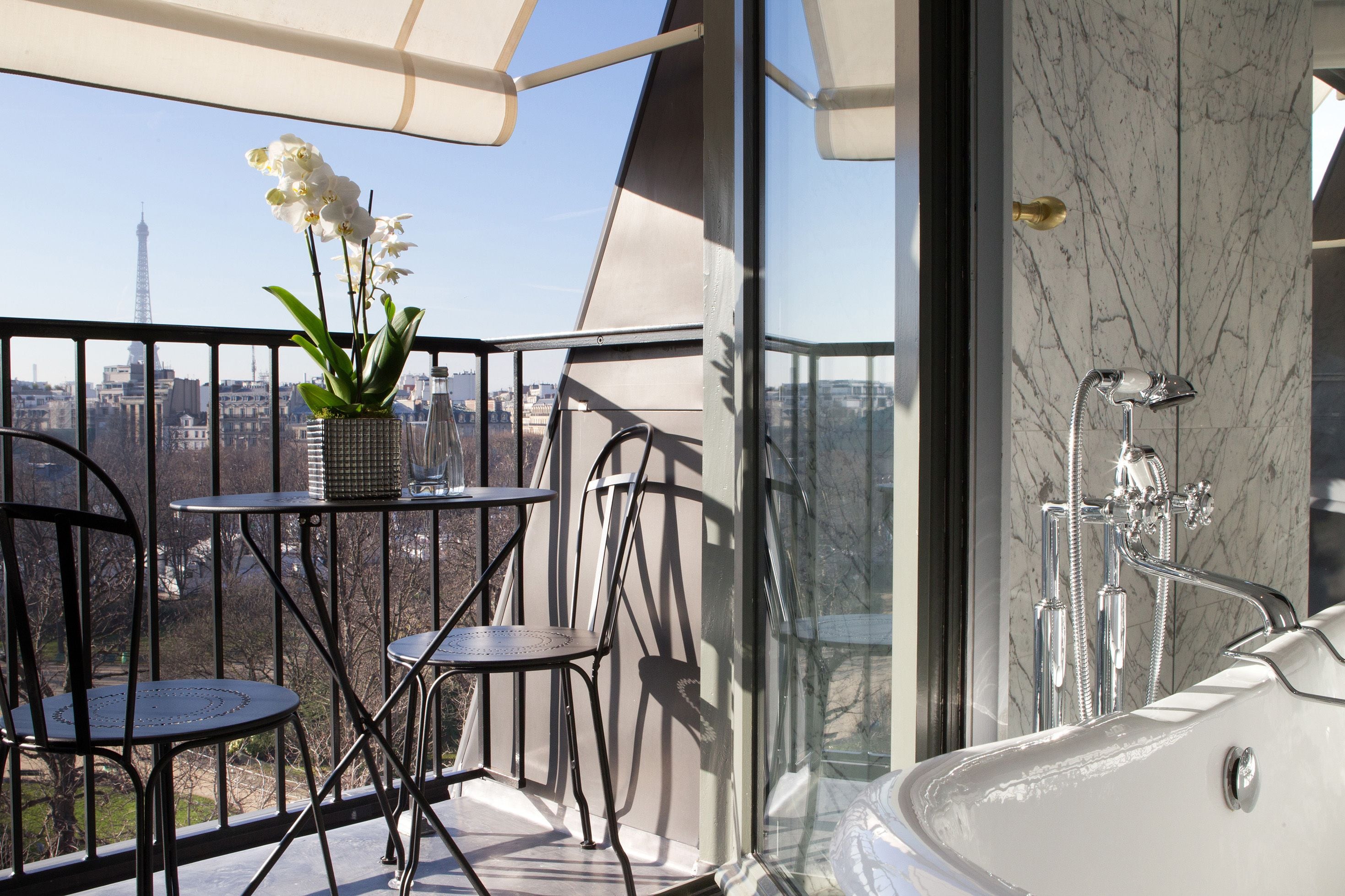 Un balcón con vistas a la Torre Eiffel, un baño con paredes de vidrio y bañera, y dos sillas.