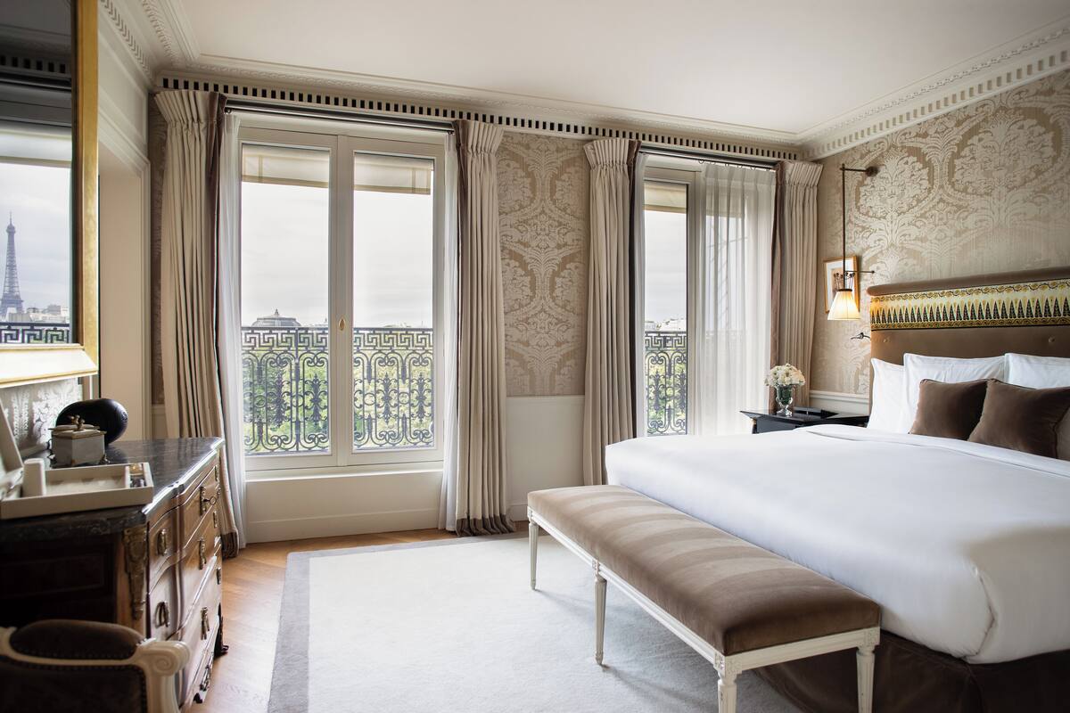 A hotel room with a large bed, a bench, a dresser, and a view of the Eiffel Tower through large windows.