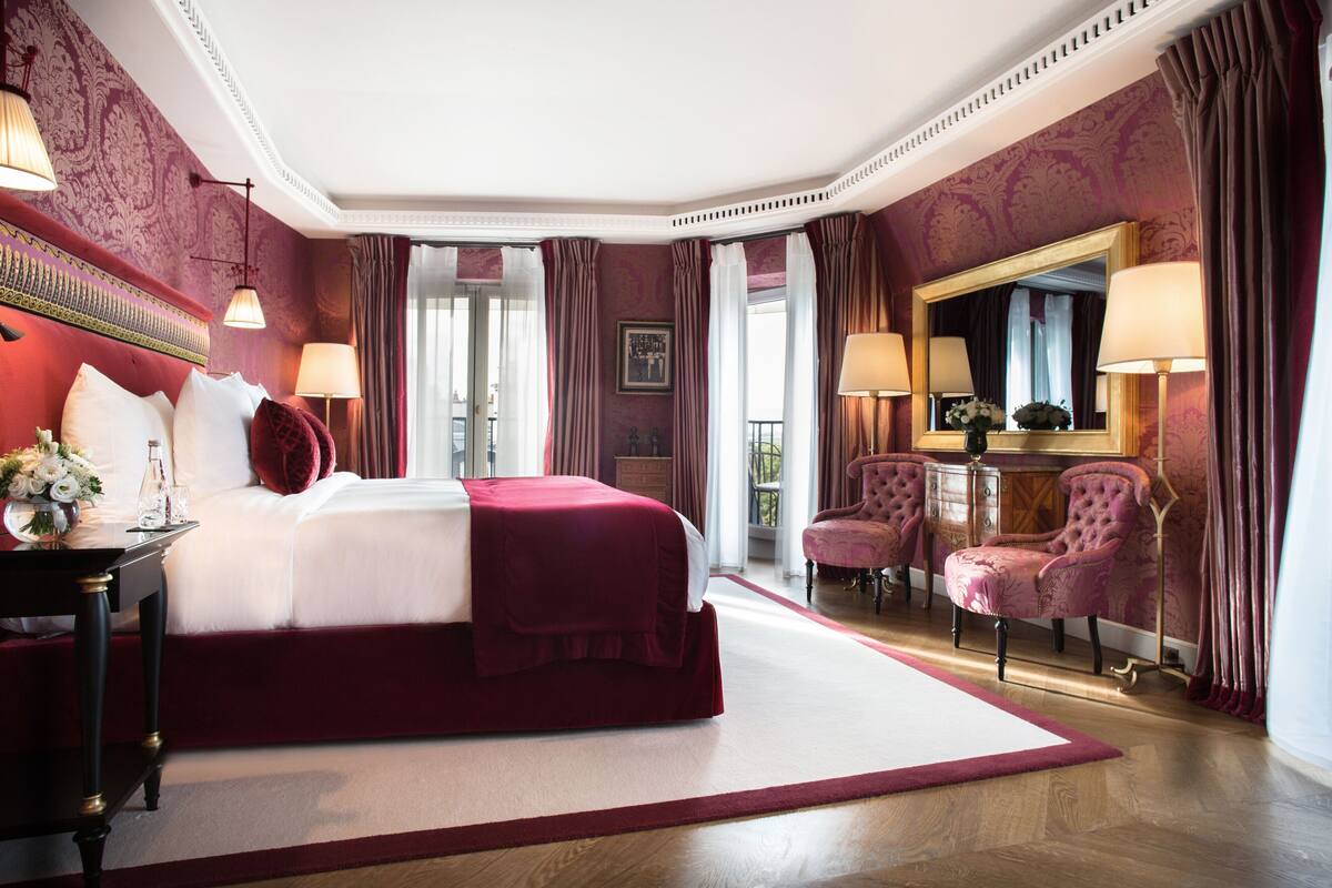 A luxurious bedroom with a large bed, two armchairs, a vanity with a mirror, and a view of the outdoors.
