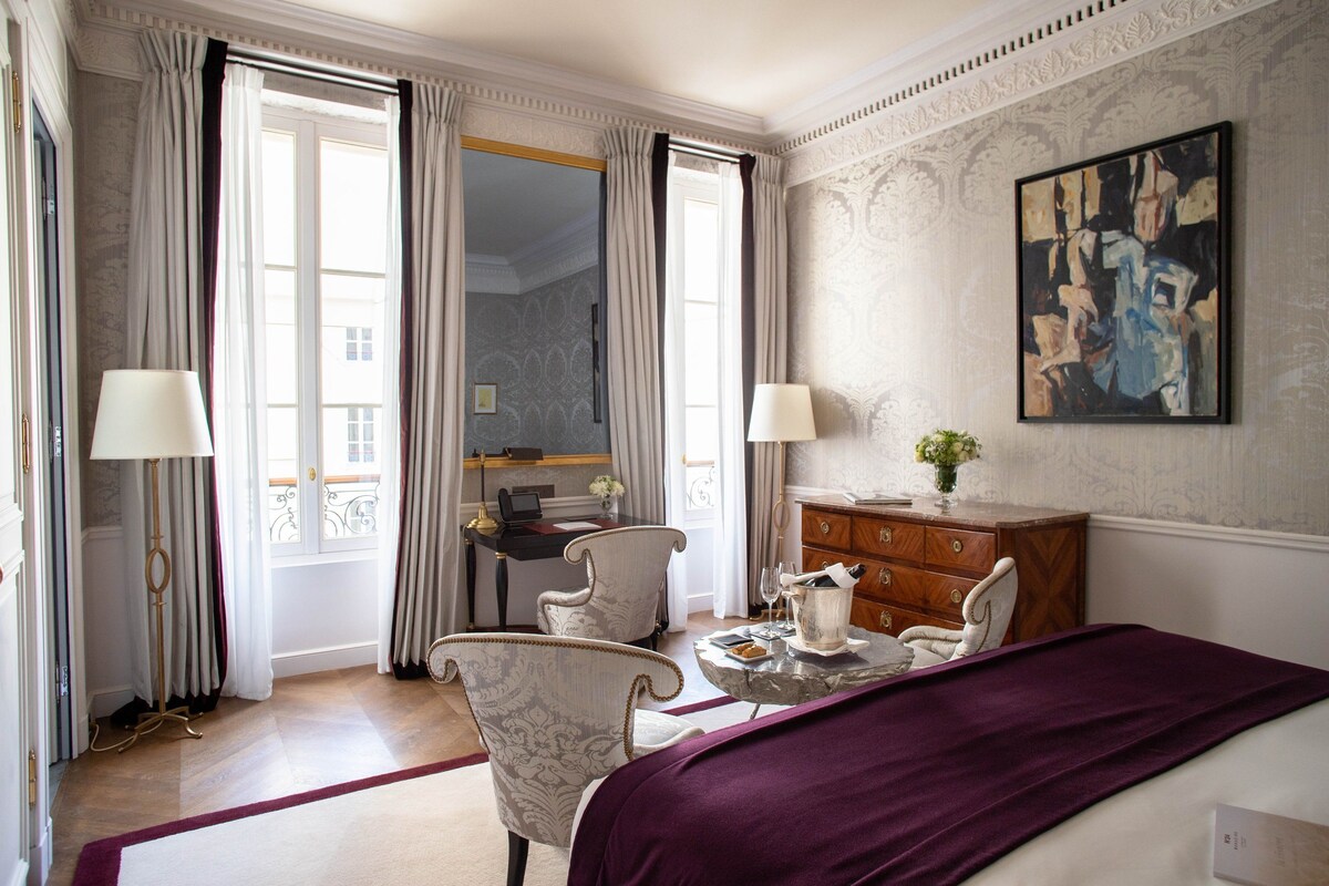 A hotel room with a bed, two chairs, a table with a vase, a dresser, and a painting on the wall.