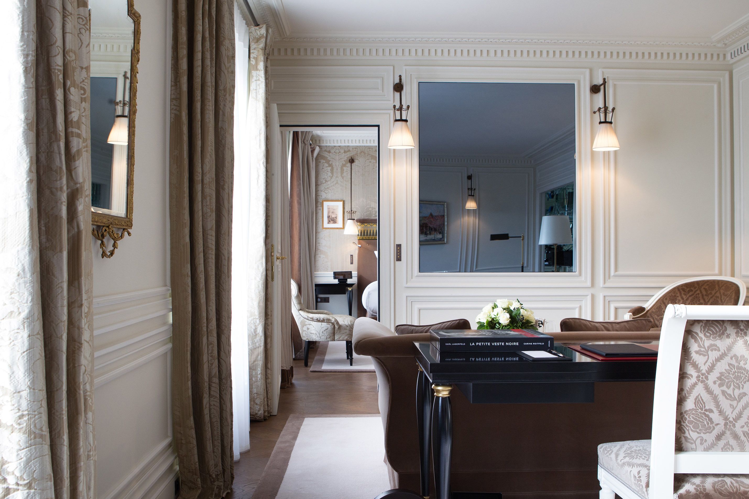 A hotel room with a large mirror, a sofa, a desk with a lamp, and a view into the adjacent room.