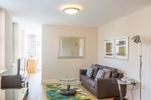 A modern living room with a brown sofa, glass coffee table, and a TV. There is a wooden side table and a patterned rug.