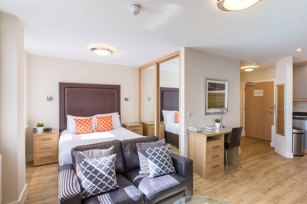 A modern hotel room with a large bed, a sofa, a desk, and a mirror.