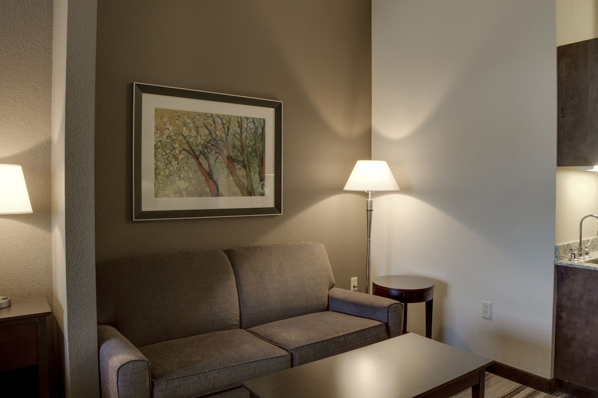 A modern hotel room with a sofa, a table, a lamp, a painting on the wall, and a small kitchen area.