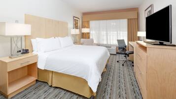 Habitación de hotel con una cama grande, dos mesitas de noche con lámparas, un escritorio con televisor de pantalla plana, una silla y un ventanal con cortinas.