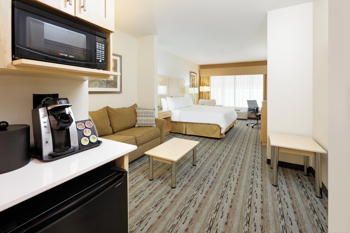 A hotel room with a kitchenette, a bed, a sofa, a desk, and a chair.
