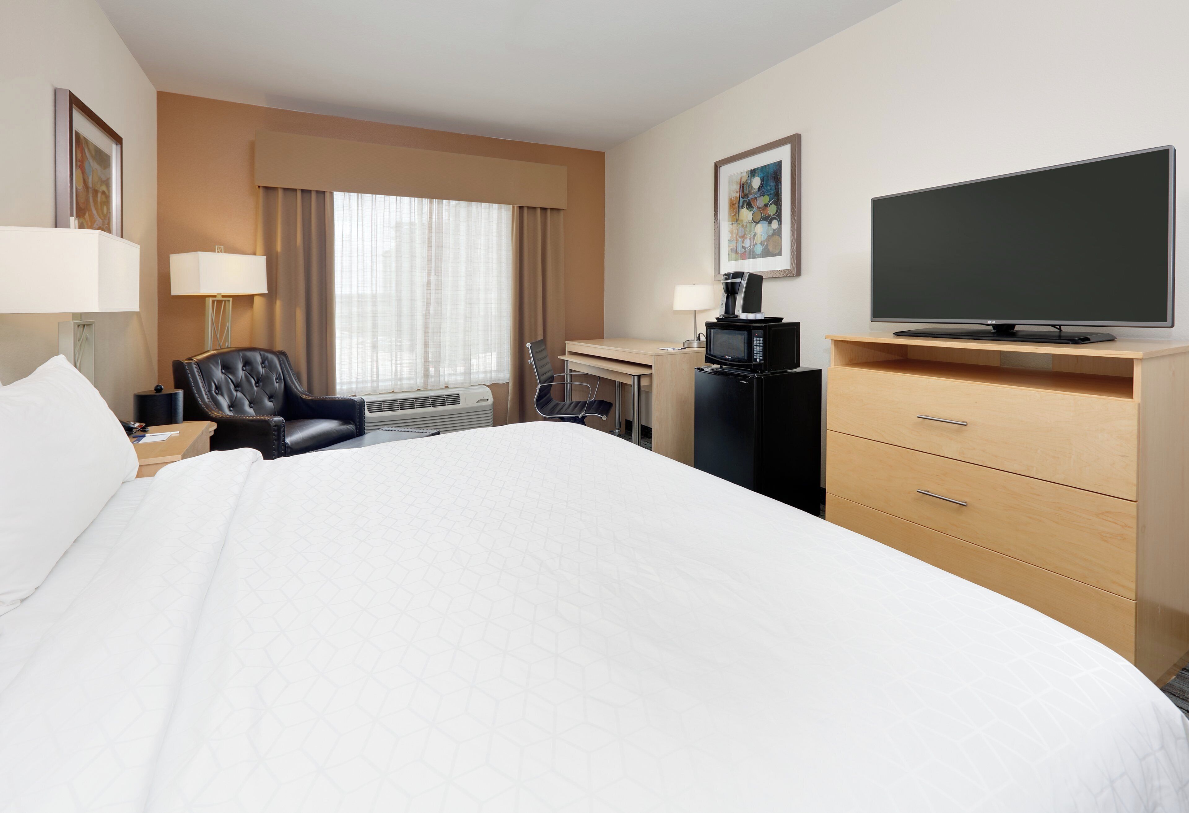 Ein Hotelzimmer mit einem großen Bett, einem Schreibtisch mit Computer, einem Fernseher, einem Sessel und einer Lampe.