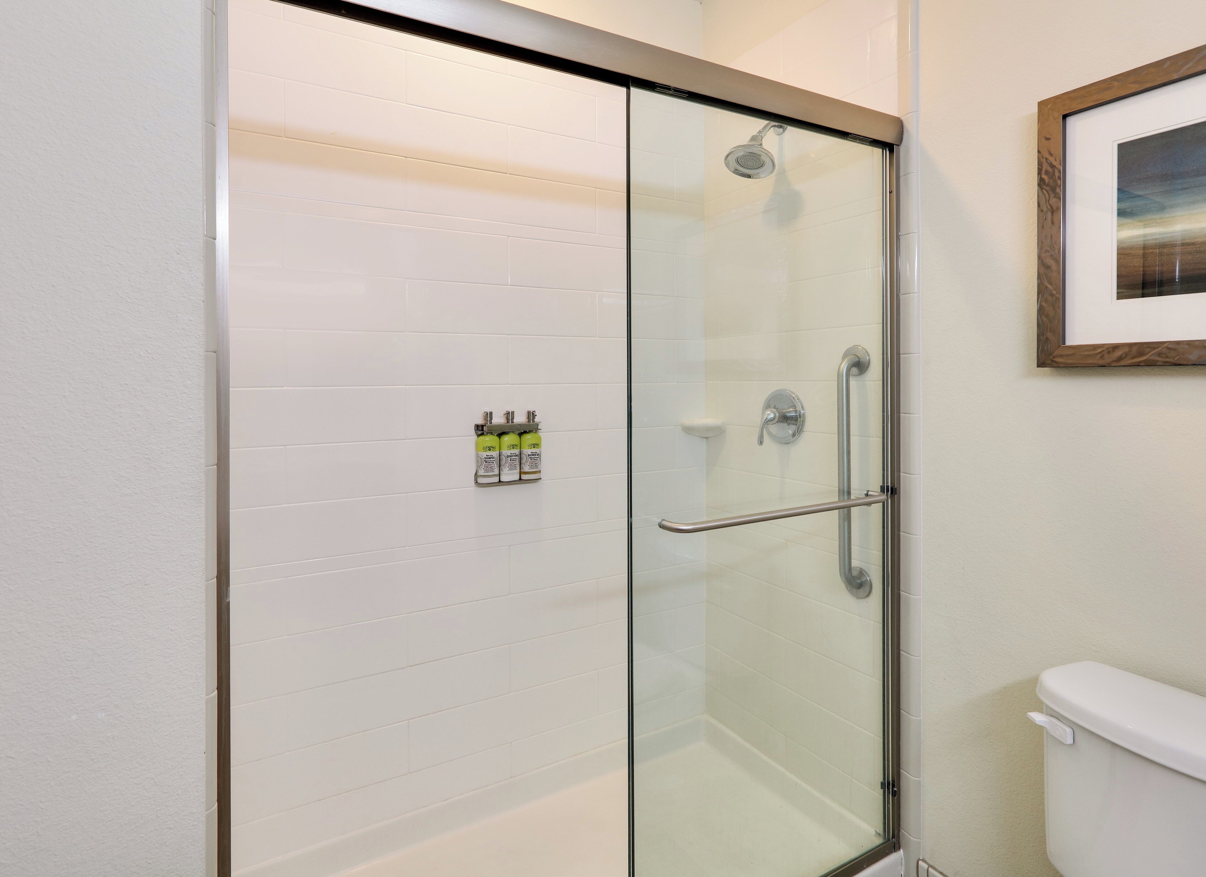 A glass-enclosed shower with a handrail, a white tiled wall, and a framed picture above the toilet.