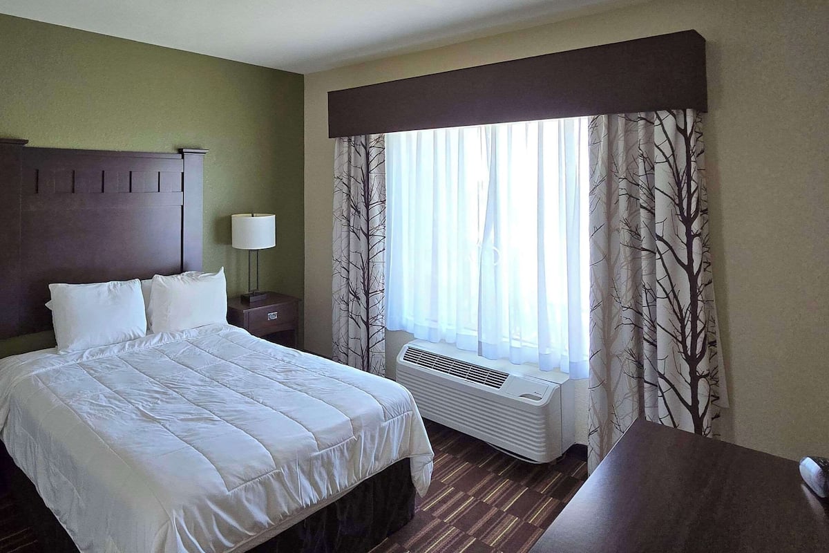 Une chambre d’hôtel comprenant un lit, deux tables de chevet avec des lampes, une fenêtre avec des rideaux légers et un climatiseur fixé au mur.