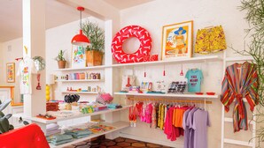 Ein hell erleuchtetes Boutique-Hotelzimmer mit einem roten Sessel, einer roten Lampe, einem Blumentopf und verschiedenen farbenfrohen Kleidungsstücken, die auf Regalen und Kleiderbügeln präsentiert werden.