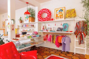 A brightly lit boutique hotel room with a red chair, a red lamp, a potted plant, and a variety of colorful clothing items displayed on shelves and hangers.