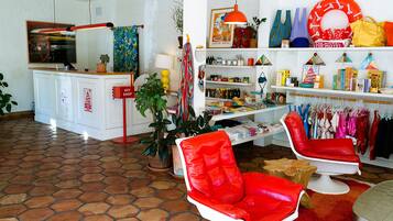 A hotel room with a tiled floor, a red chair, a white chair, a desk with a computer, shelves with various items, and a plant.