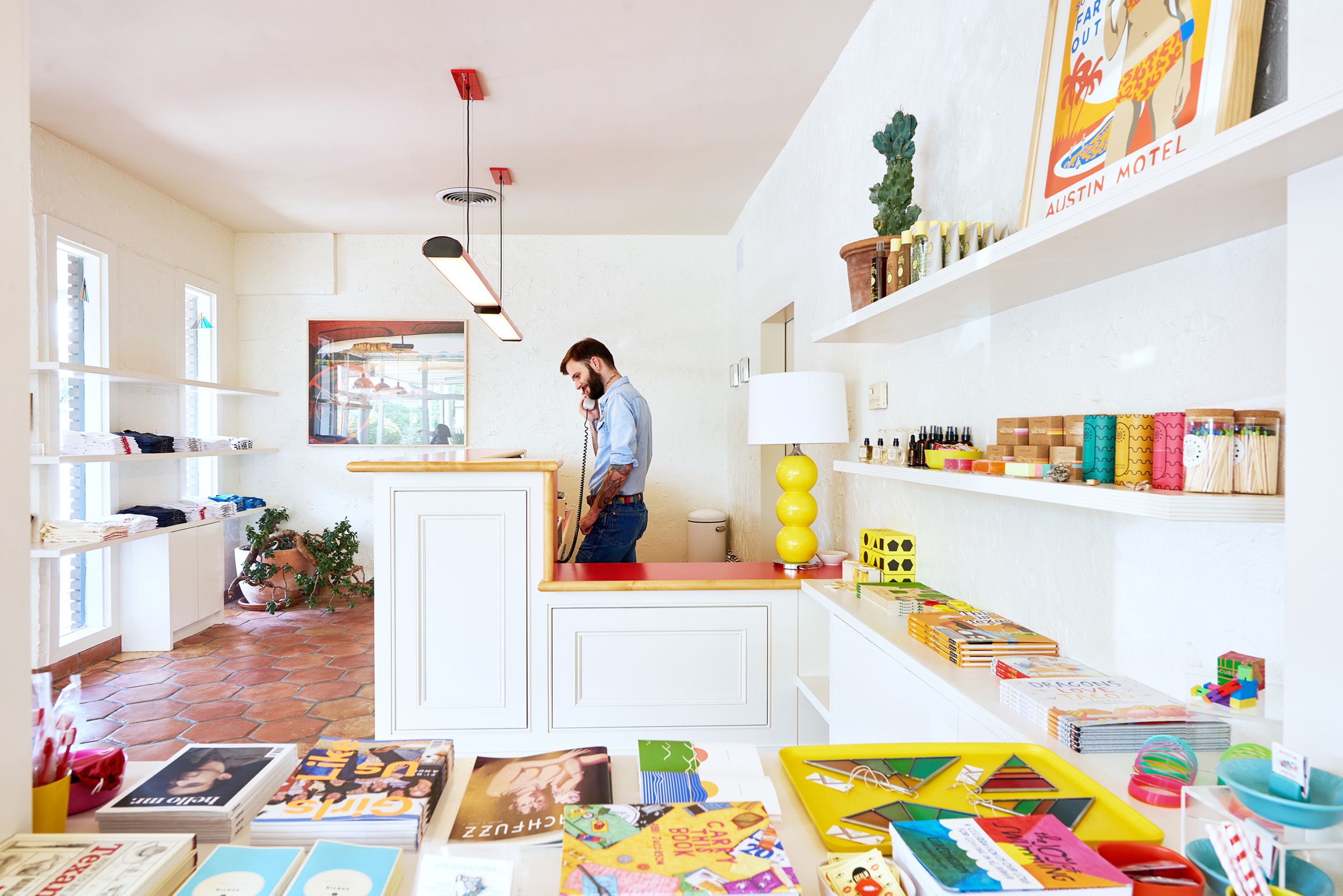 Ein helles Zimmer mit Schreibtisch, Regalen mit Büchern und Pflegeprodukten sowie einem Blumentopf.