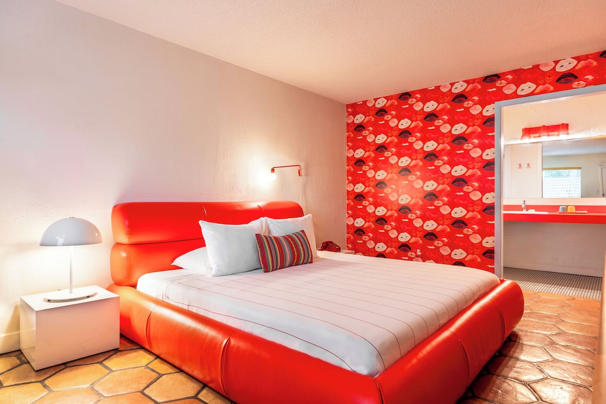 A modern hotel room with a red upholstered bed, a white nightstand, a striped pillow, and a red patterned wall.