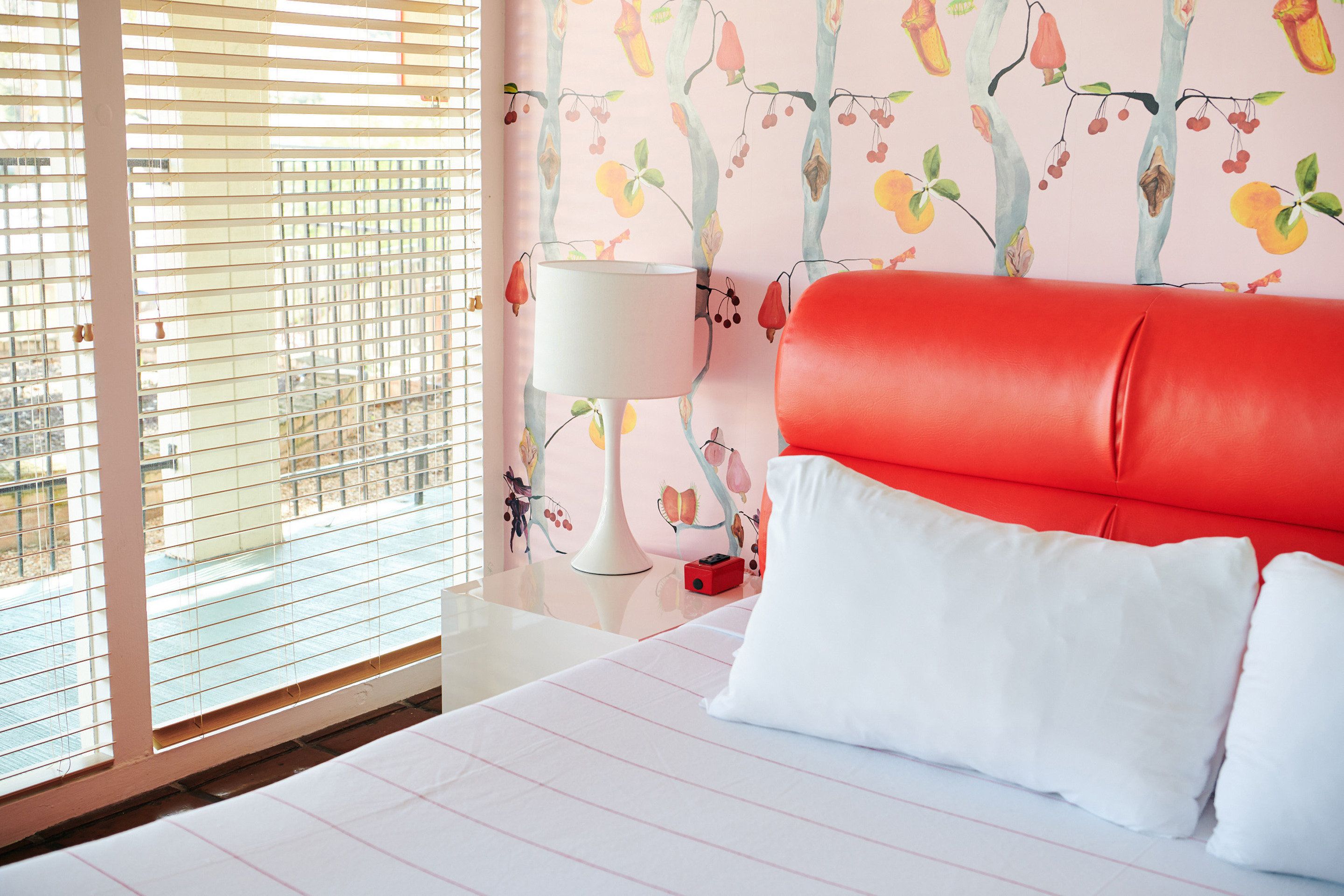 Une chambre d’hôtel avec un lit, une tête de lit rouge, une lampe blanche et une fenêtre avec des stores.