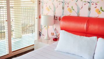 Une chambre d’hôtel avec un lit, une tête de lit rouge, une lampe blanche et une fenêtre avec des stores.