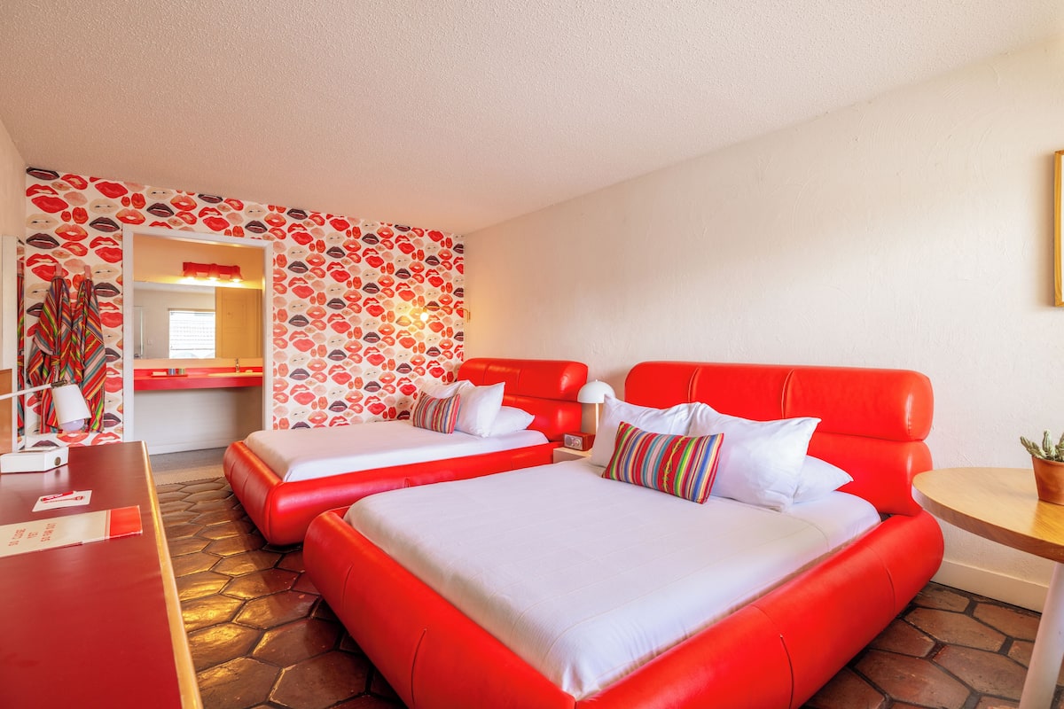 A hotel room with two red beds, white bedding, a red sofa, a small table with a plant, and a wall with a red and white pattern.