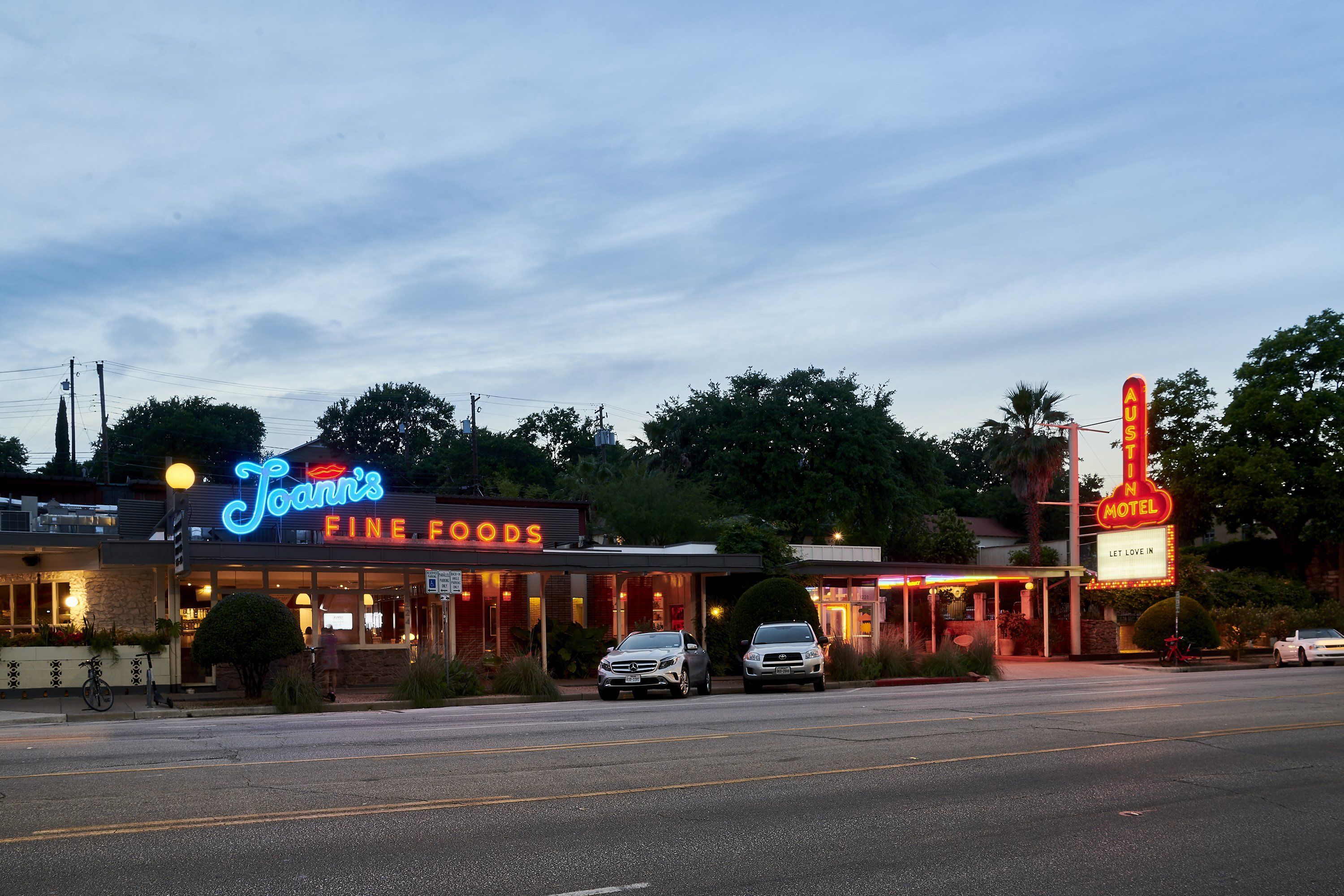 Un motel con letreros de neón, un restaurante llamado "Joanni's Fine Foods" y varios autos estacionados.