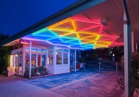 A motel with a neon-lit geometric ceiling, a 'VACANCY' sign, and a 'YALL MEANS ALL!' banner.