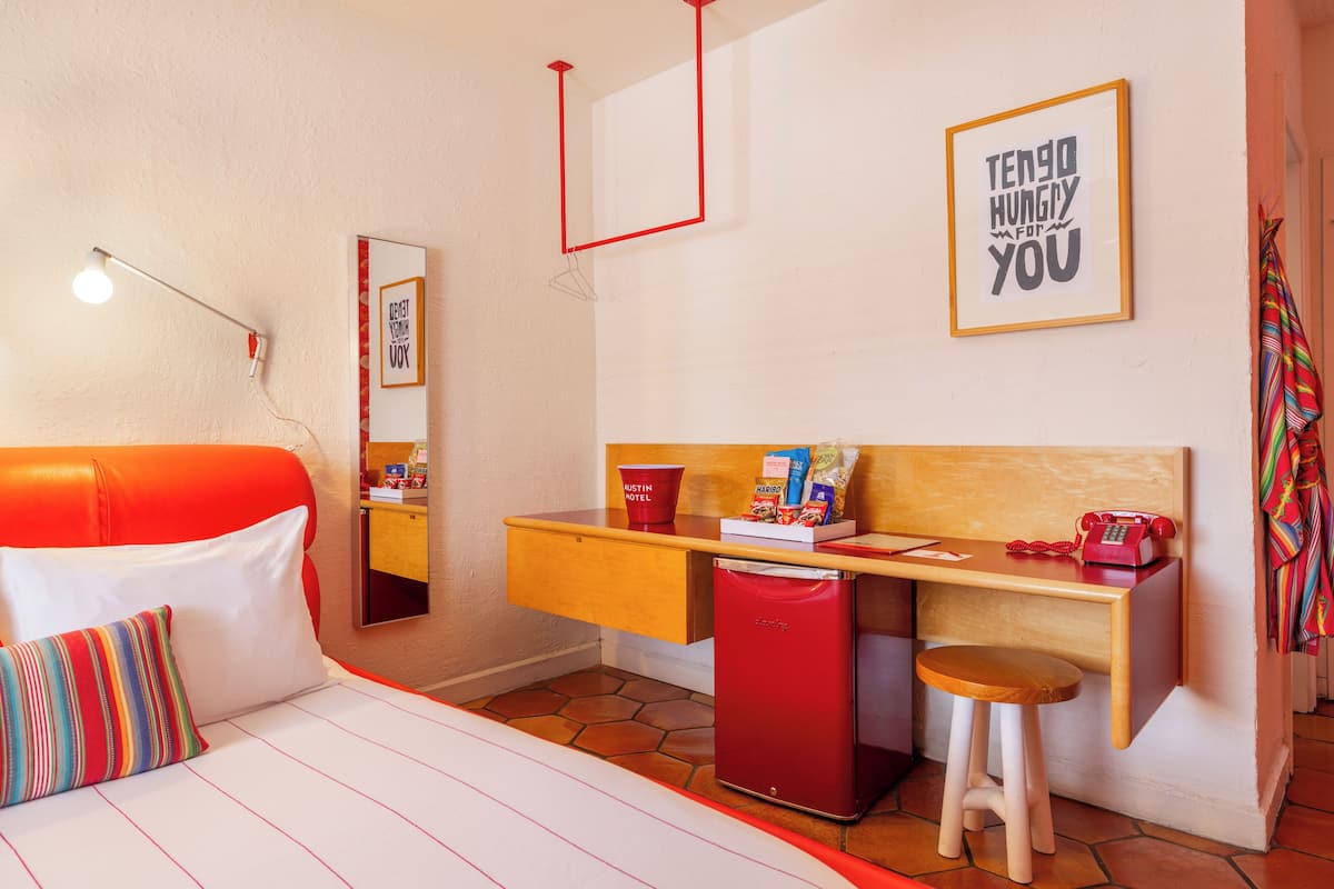 A compact hotel room with a bed, a desk, a small refrigerator, and a stool.