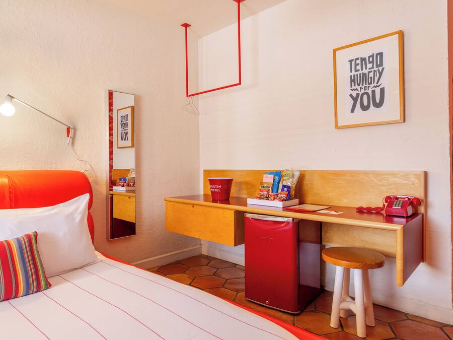 A compact hotel room with a bed, a desk, a small refrigerator, and a stool.