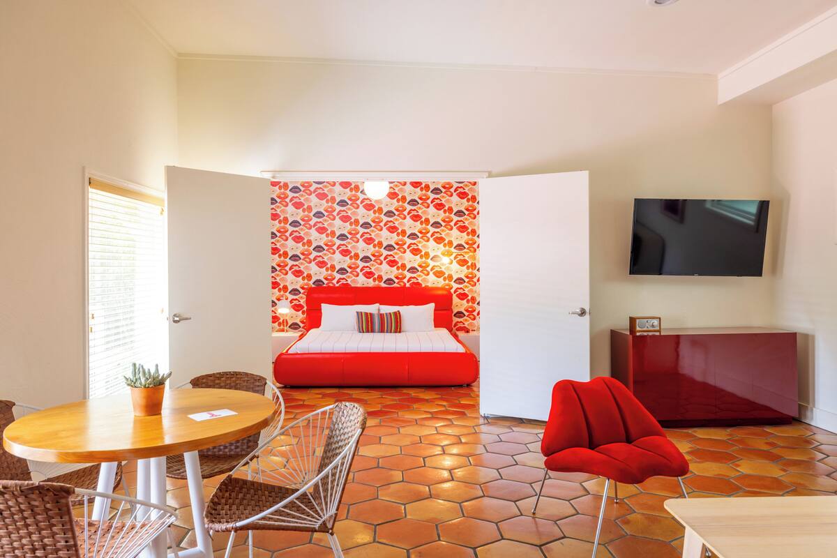 A modern hotel room with a red bed, a television, a dining area with wicker chairs, and a tiled floor.
