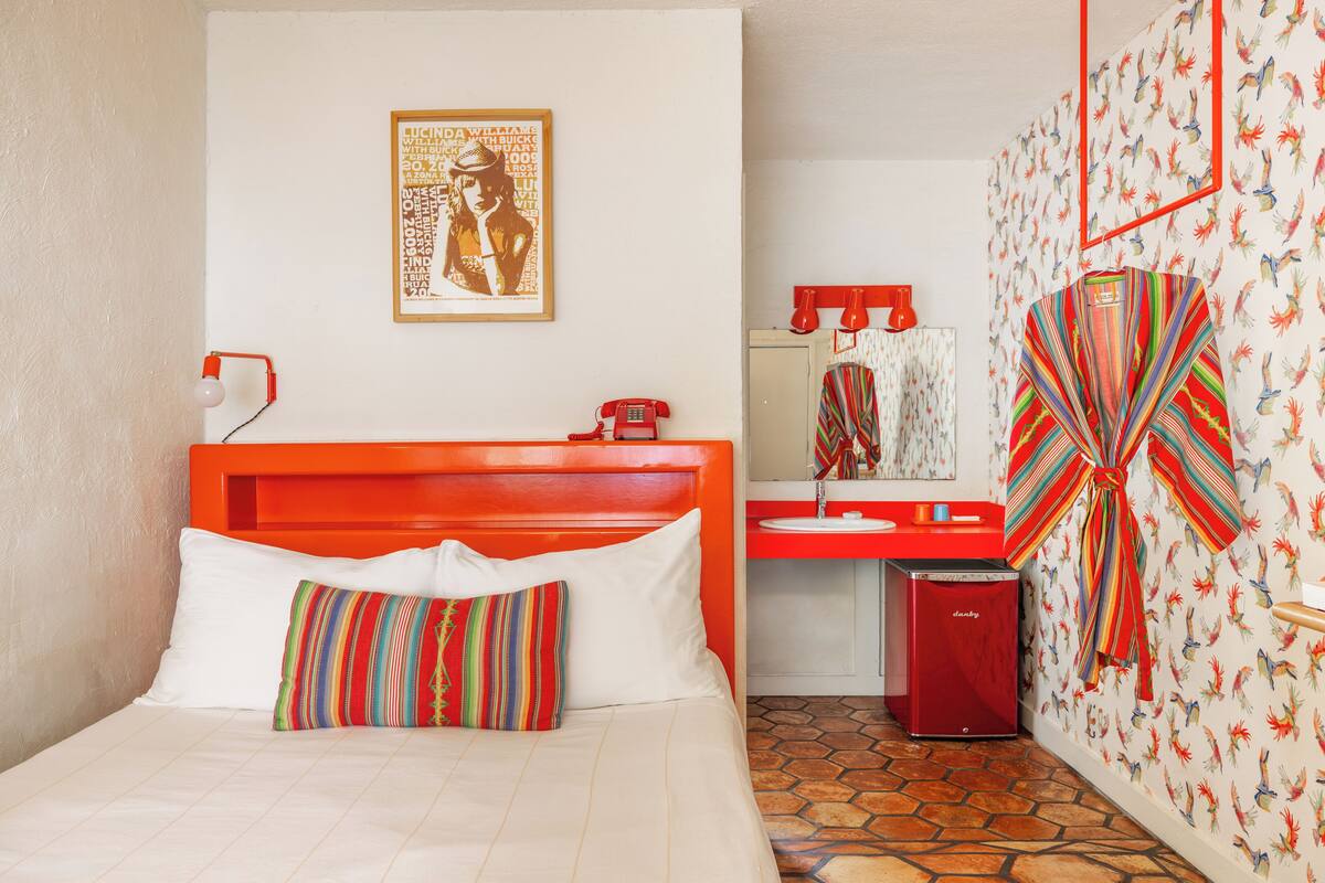 A hotel room with a bed, a red telephone, a red minibar, a mirror, and a wall with colorful bird wallpaper.