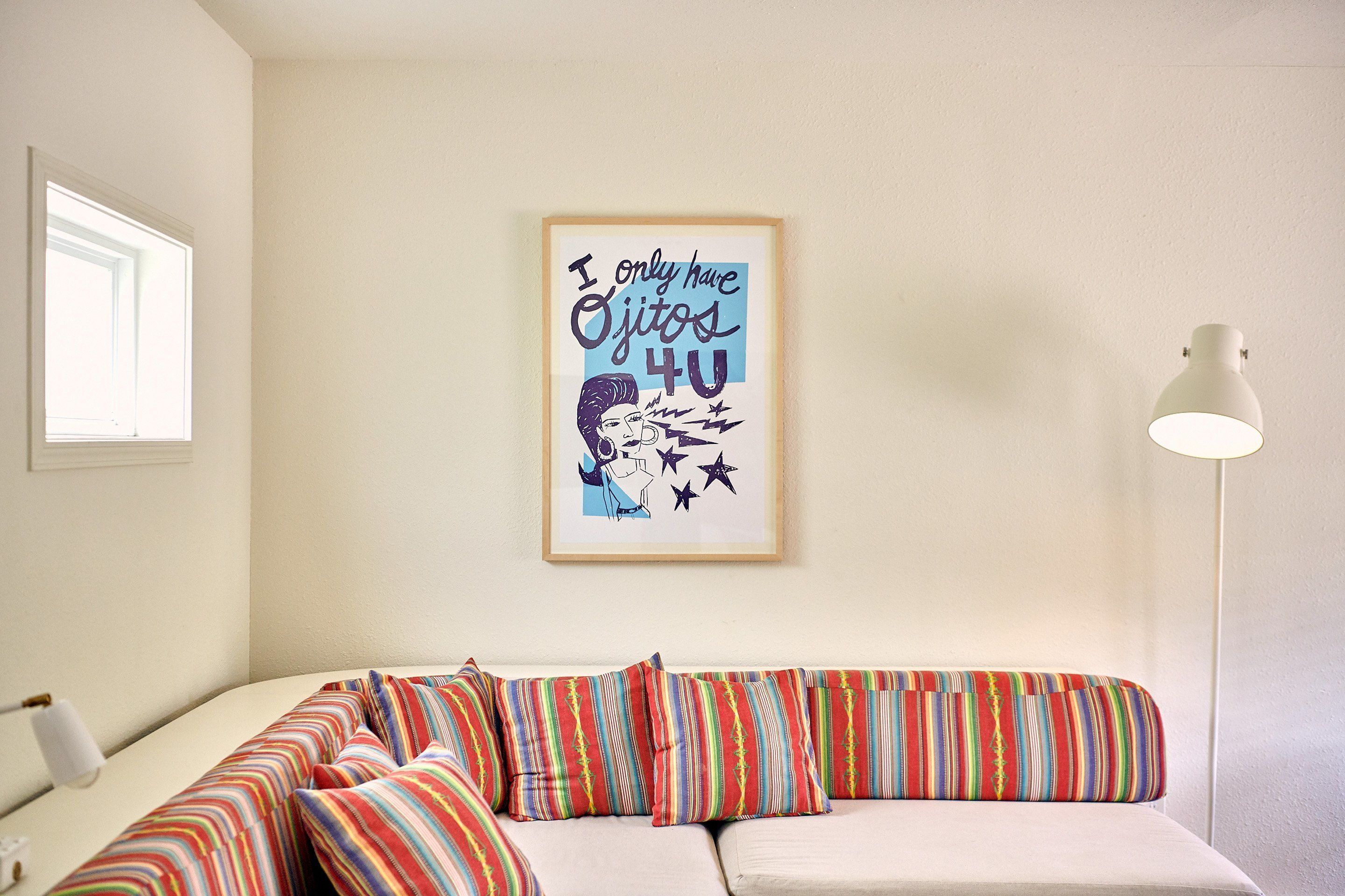 Una habitación con un sofá blanco, almohadones a rayas de colores, un cartel enmarcado en la pared y una lámpara de pie blanca.