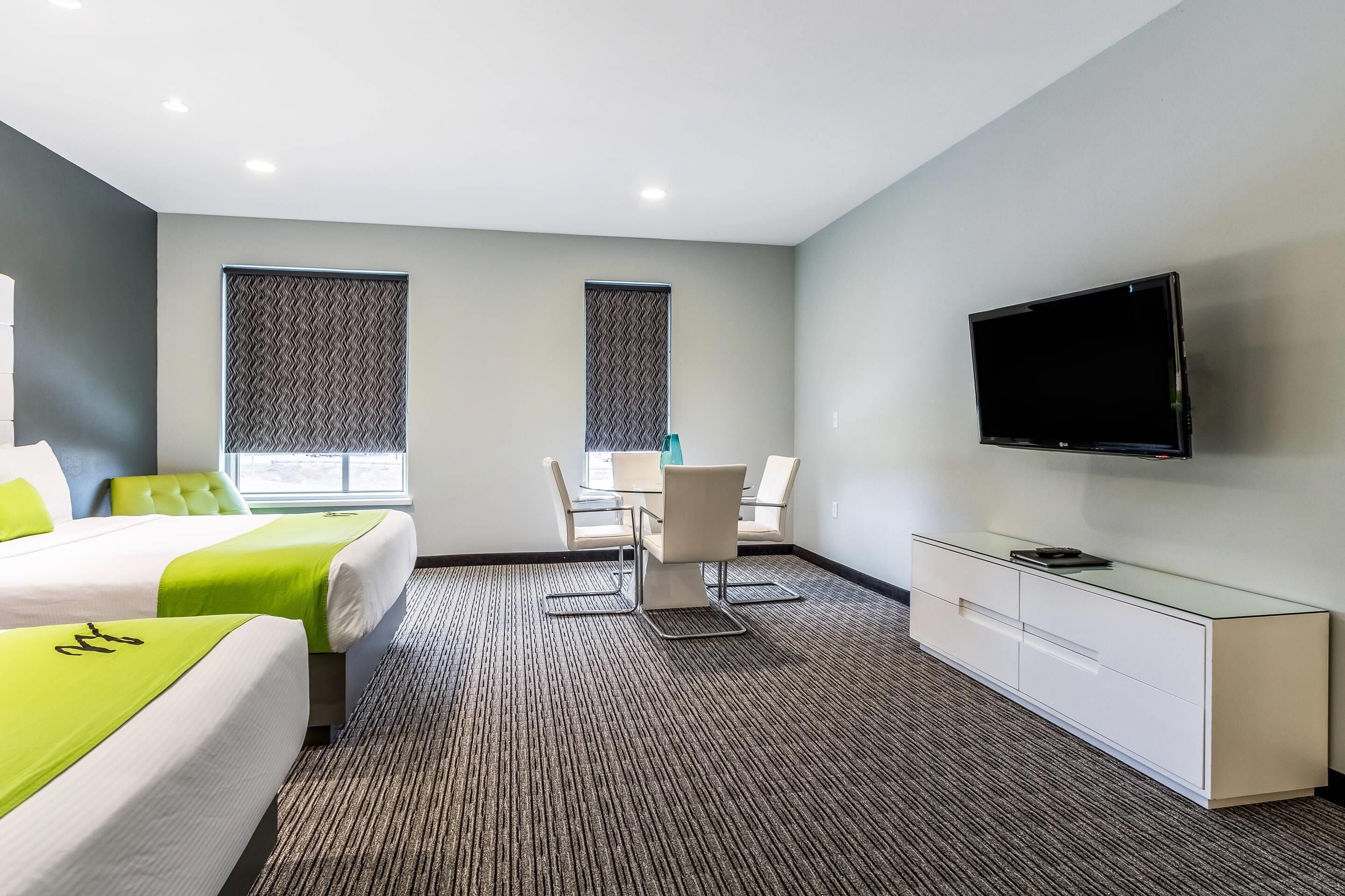 A hotel room with two beds, a dining table with chairs, a TV on the wall, and a carpeted floor.