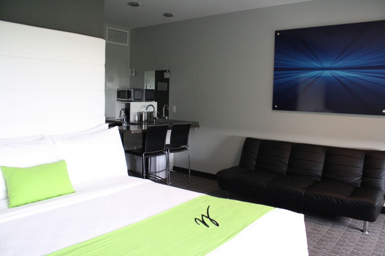 A hotel room with a bed, a green throw, a black leather sofa, a dining area with a kitchen, and a large abstract painting on the wall.