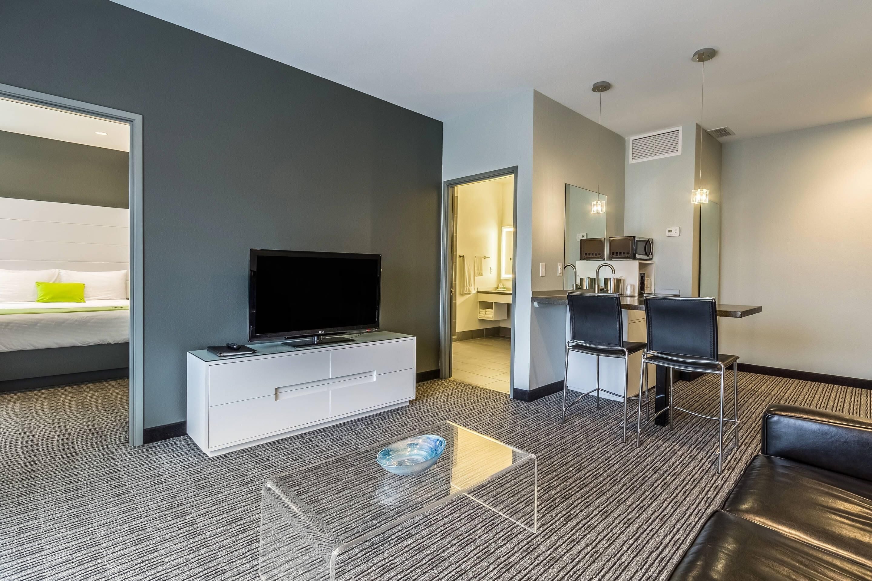 A modern hotel room with a flat-screen TV, a white console, a glass coffee table, a black leather sofa, a dining area with chairs, and a kitchen area with a microwave.