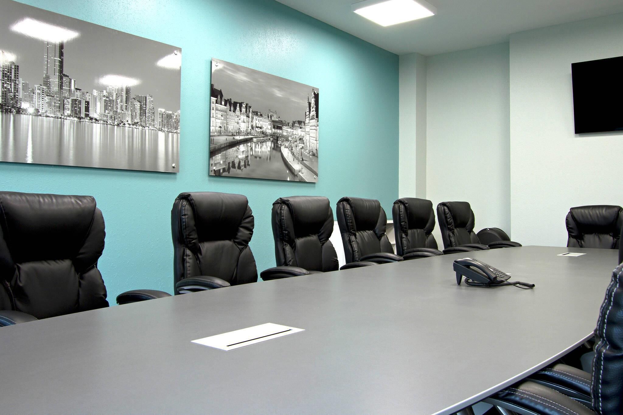 A conference room with a long table, six black leather chairs, two black and white photographs on the wall, and a flat-screen TV mounted on the wall.