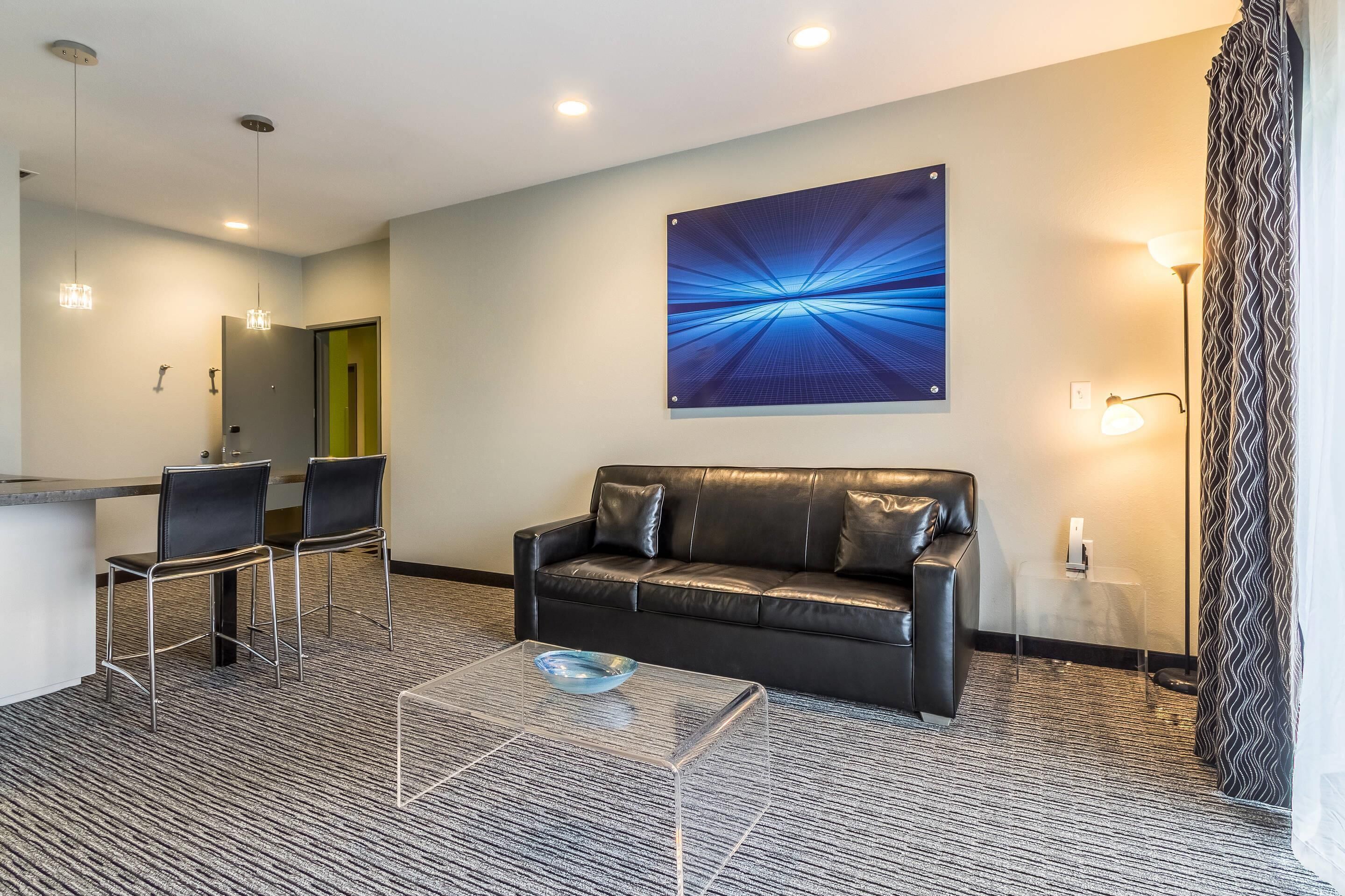 A hotel room with a black leather sofa, two chairs, a glass coffee table, a painting on the wall, a desk with a chair, and a patterned carpet.