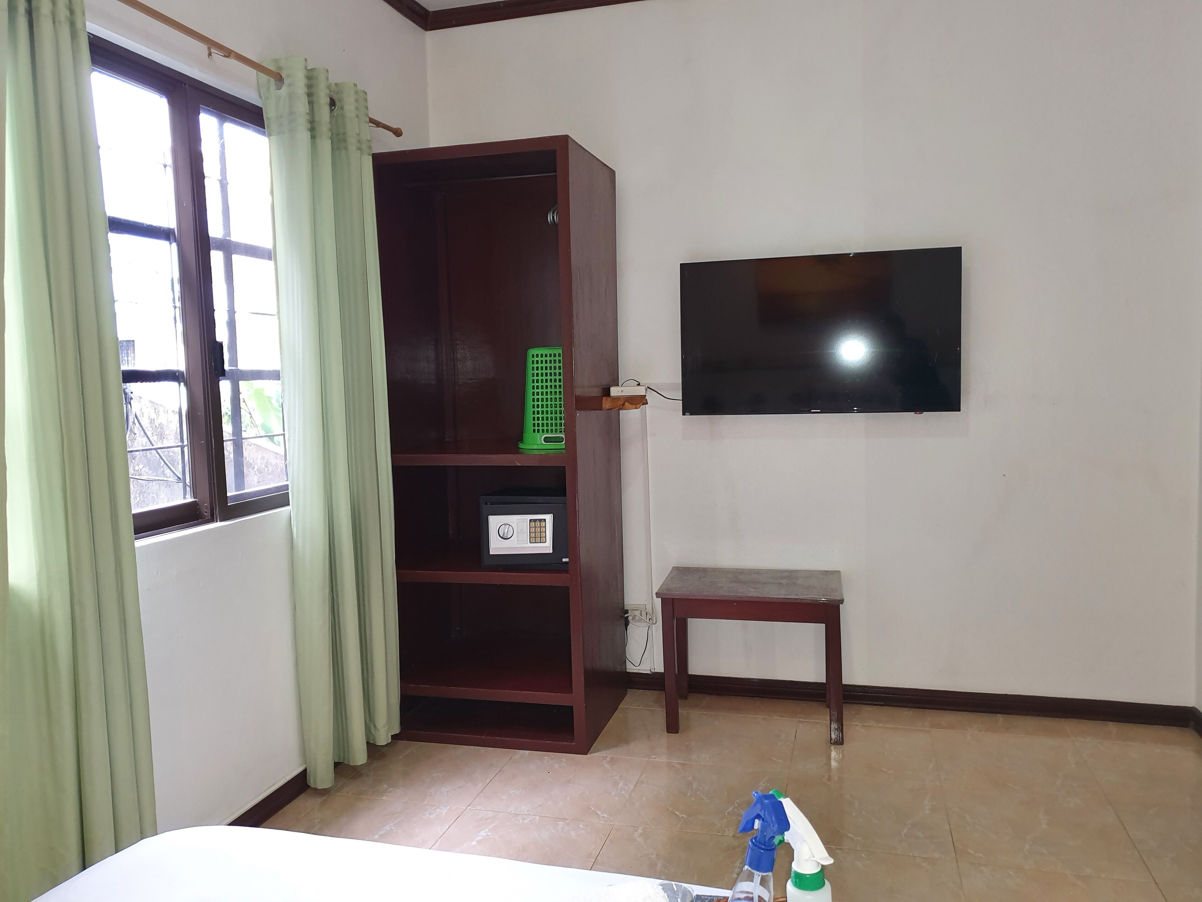 A room with a TV mounted on the wall, a wooden cabinet, a small table, and a window with curtains.