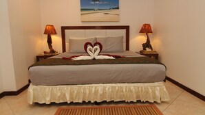Una cama impecablemente arreglada con una toalla dispuesta en forma de corazón, dos lámparas de noche y un cuadro con motivos playeros en la pared.