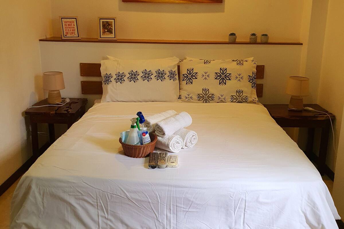 Una habitación de hotel con cama, almohadas, una mesita de noche, una lámpara, un cuadro de un atardecer y un estante con adornos.