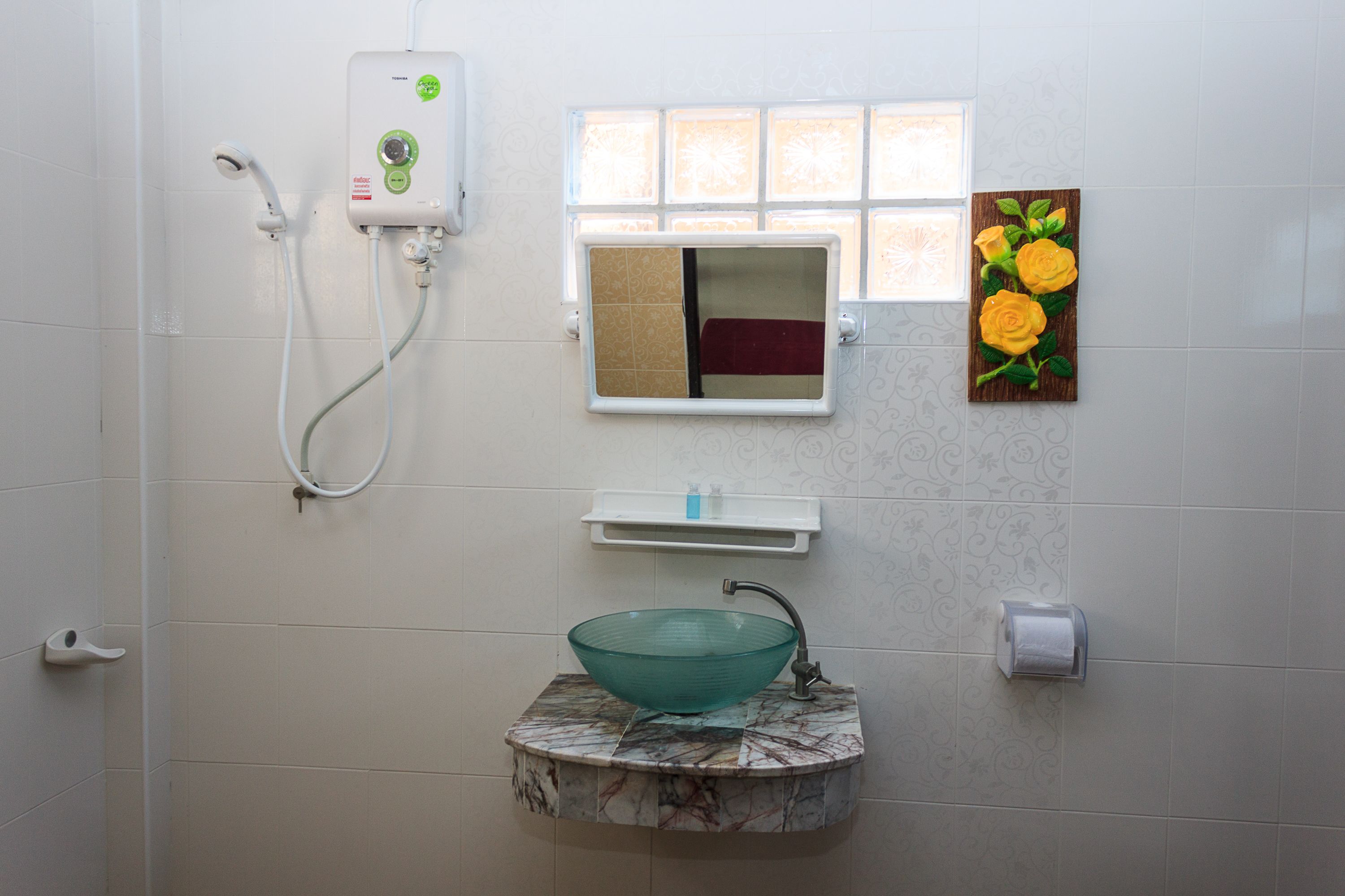 Un baño con un lavabo de vidrio, un cabezal de ducha de mano, un ventanal con espejo y un dispensador de papel higiénico en la pared.