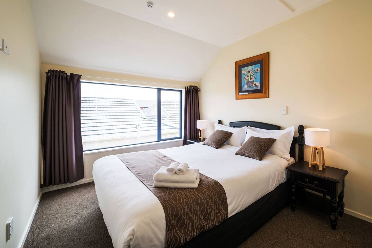 A neatly made bed with a brown accent, a bedside table with a lamp, and a window with curtains.