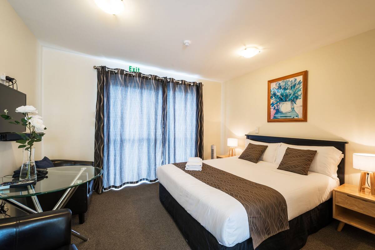 A hotel room with a bed, a desk with a chair, a glass table, a lamp, and a painting on the wall.